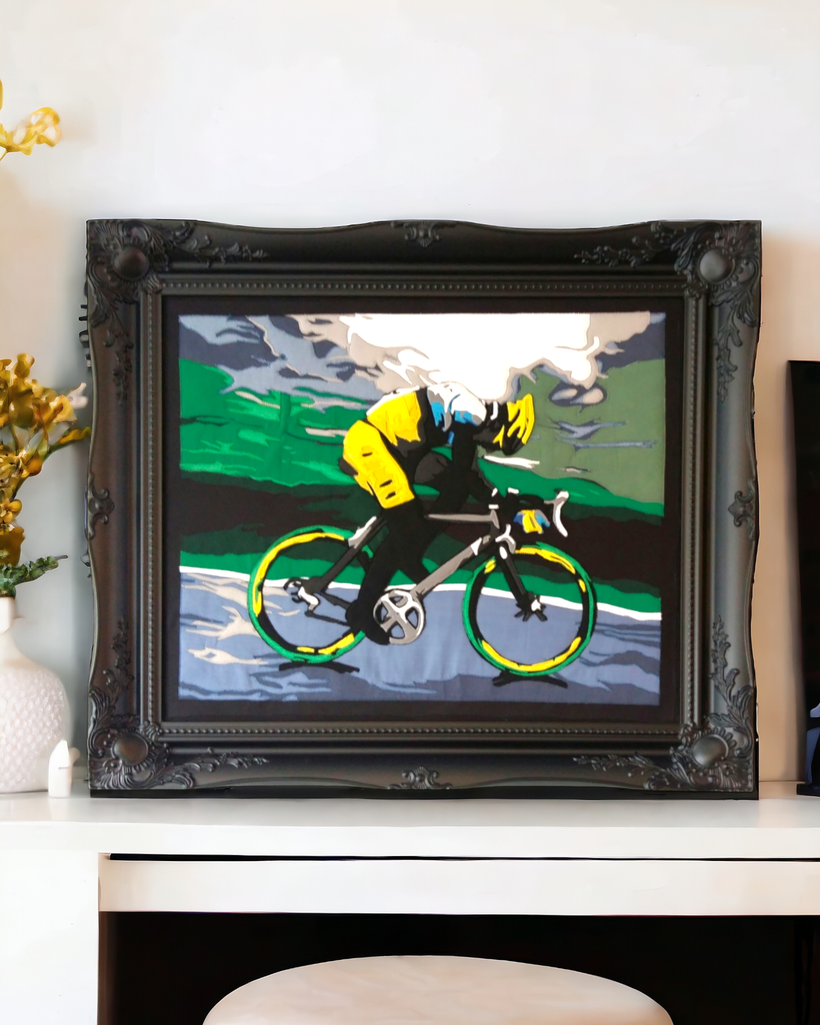 Applique fabric art peice of a cyclist riding along on a wet, rainy day in the uk. Finish in a black Swept framed with white livingroom decor in the background.