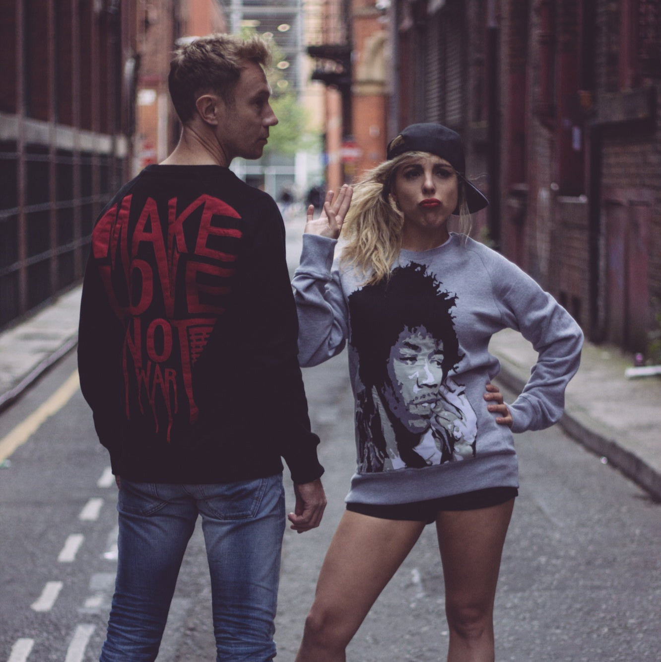 Male and Female models in One of a Kind Custom Appliqued Sweatshirts, Make Love Not War & 4 Layers of fabric in a Jimi Hendricks silhouette, cut and sewed onto a black and grey sweatshirts. Street ally of manchester fashion quater in the background.